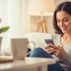 Uśmiechnięta kobieta na kanapie przegląda telefon, szukając sposobu, jak kogoś rozśmieszyć do łez.
