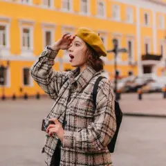 Dziewczyna w żółtym beracie i kratowej kurtce z aparatem szuka inspiracji na ciekawe tematy do rozmowy z dziewczyną.