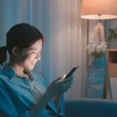 Uśmiechnięta dziewczyna pisze SMS-a "myślę o tobie" na telefonie, siedząc wieczorem w przytulnym pokoju.