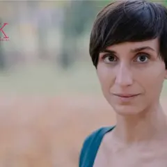 Kobieta po przejściach, z krótkimi włosami i przenikliwym spojrzeniem, uśmiecha się lekko. W tle logo "20 lat Miasto Kobiet".