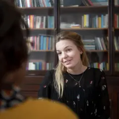 Kobieta z blond włosami, w czarnej bluzce, rozmawia z kimś, kto siedzi tyłem. Za nią regały z książkami. Może to być przykład odwróconej psychologii w związku.