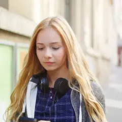 Dziewczyna z blond włosami i słuchawkami na szyi przegląda telefon, zastanawiając się, jak zacząć rozmowę z chłopakiem.