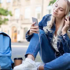 Dziewczyna w dżinsowej kurtce słucha muzyki na telefonie. Obok niej leży niebieski plecak i książki. Idealny moment, by pomyśleć, o czym można pisać z chłopakiem.