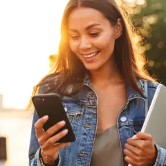 Uśmiechnięta dziewczyna w dżinsowej kurtce czyta na telefonie dwuznaczne teksty do dziewczyny, trzymając laptopa.