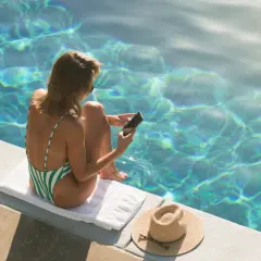 Kobieta w zielono-białym stroju kąpielowym siedzi przy basenie, trzymając telefon. Nie pisze pierwsza, ale odpisuje, ciesząc się słońcem i wodą.