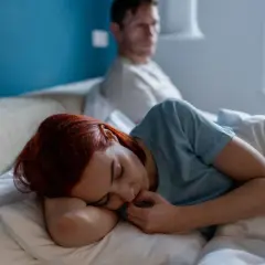 Kobieta z rudymi włosami śpi, czuje się zaniedbywana w związku. Mężczyzna siedzi z tyłu, odwrócony.