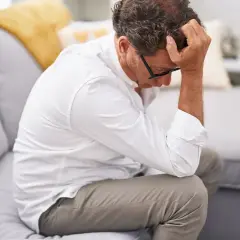 Mężczyzna siedzi na kanapie, trzymając się za głowę. Wygląda na zmartwionego, jakby właśnie usłyszał od wiecznie niezadowolonej żony, że znów zapomniał kupić mleko.