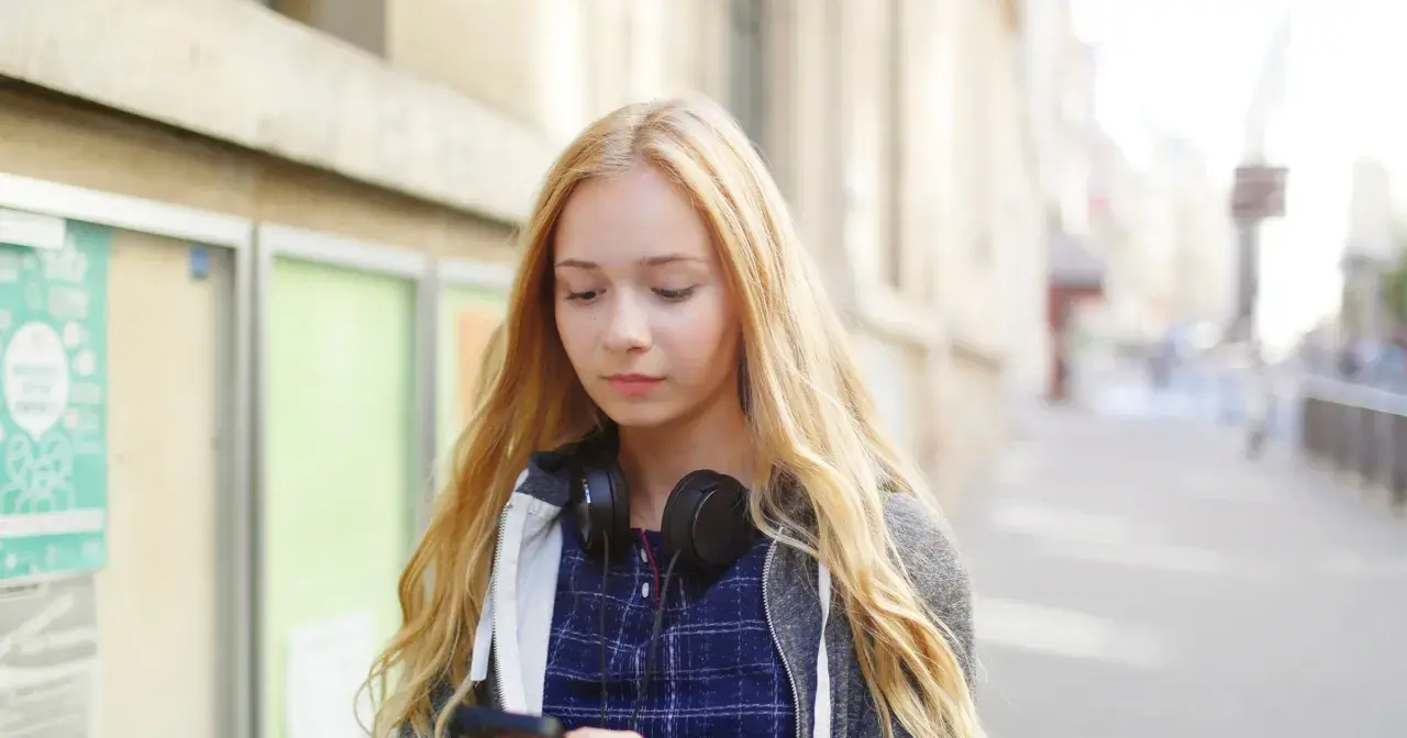 Dziewczyna z blond włosami i słuchawkami na szyi przegląda telefon, zastanawiając się, jak zacząć rozmowę z chłopakiem.