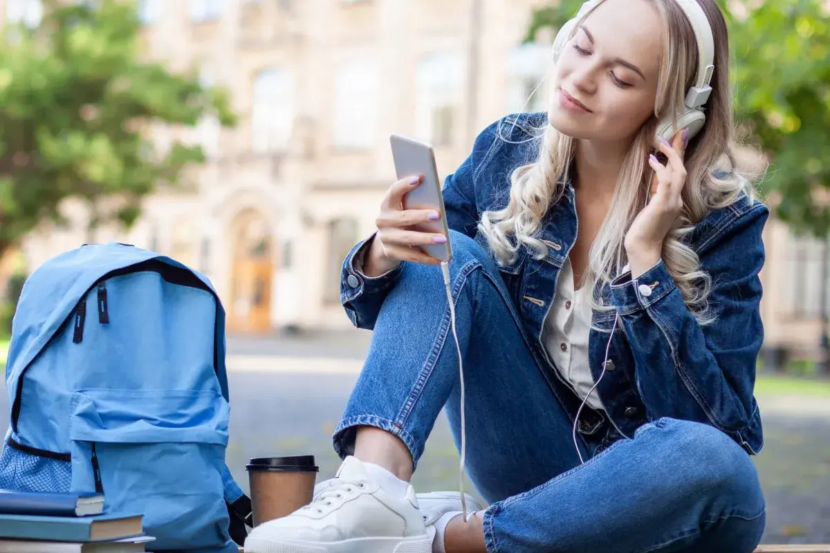 Dziewczyna w dżinsowej kurtce słucha muzyki na telefonie. Obok niej leży niebieski plecak i książki. Idealny moment, by pomyśleć, o czym można pisać z chłopakiem.