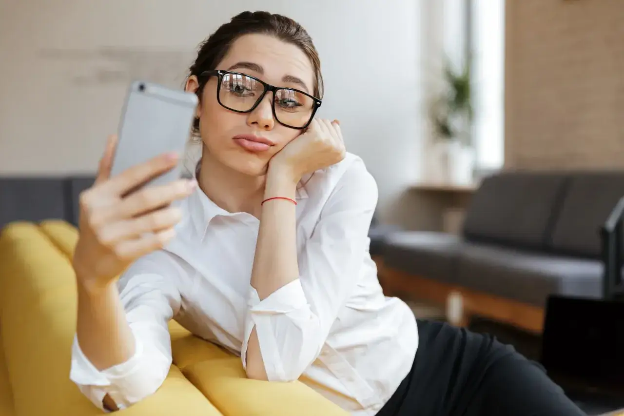 Kobieta w okularach leży na kanapie, trzymając telefon. Wyświetla a nie odpisuje, czekając na wiadomość.
