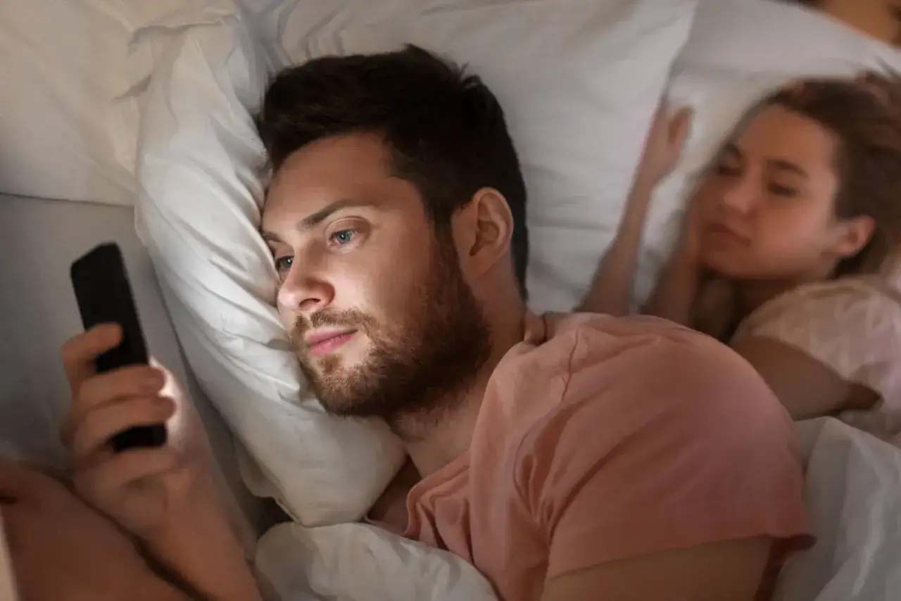 Mężczyzna w łóżku patrzy na telefon, podczas gdy jego partnerka śpi. Co oznacza gdy dziewczyna pisze dobranoc? Może to być znak, że chce zakończyć rozmowę lub potrzebuje przestrzeni.