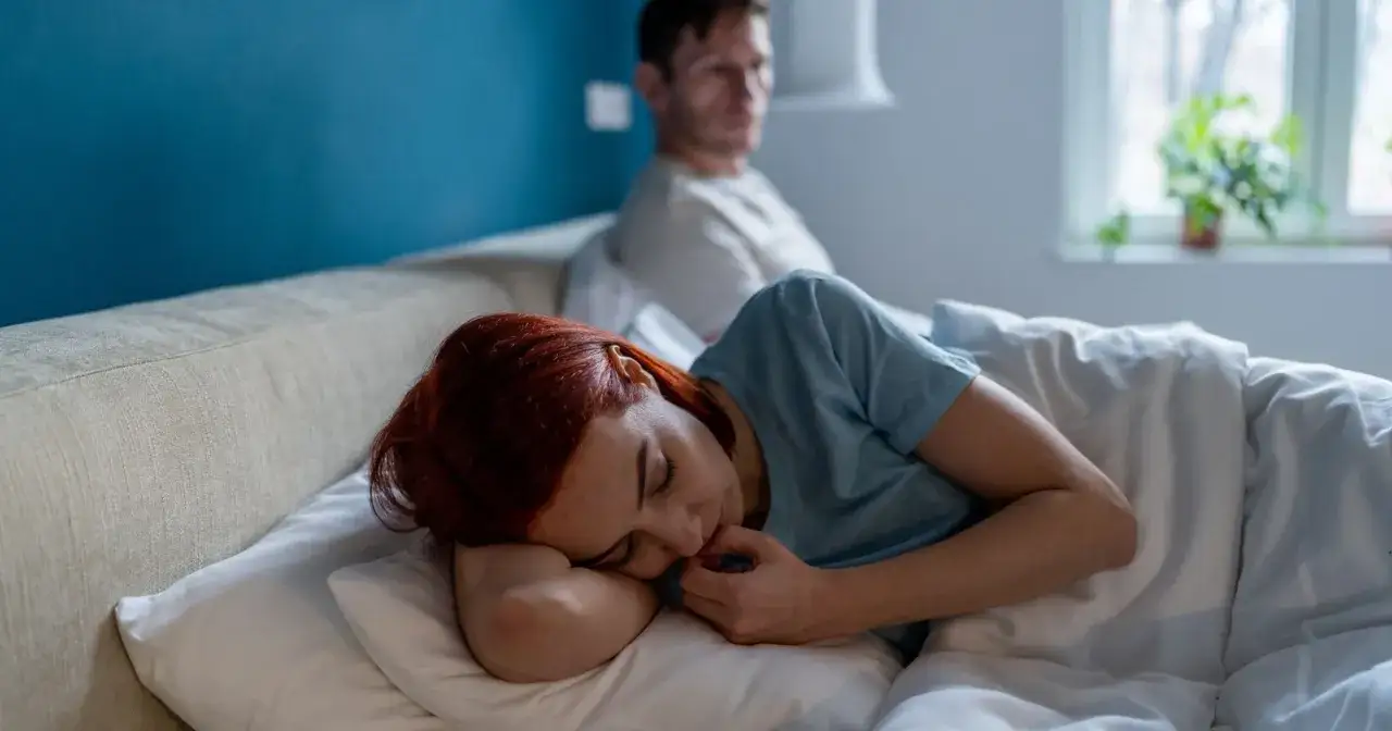 Kobieta z rudymi włosami śpi, przytulając poduszkę. Mężczyzna siedzi z tyłu, odwrócony. Czuje się zaniedbywana w związku.