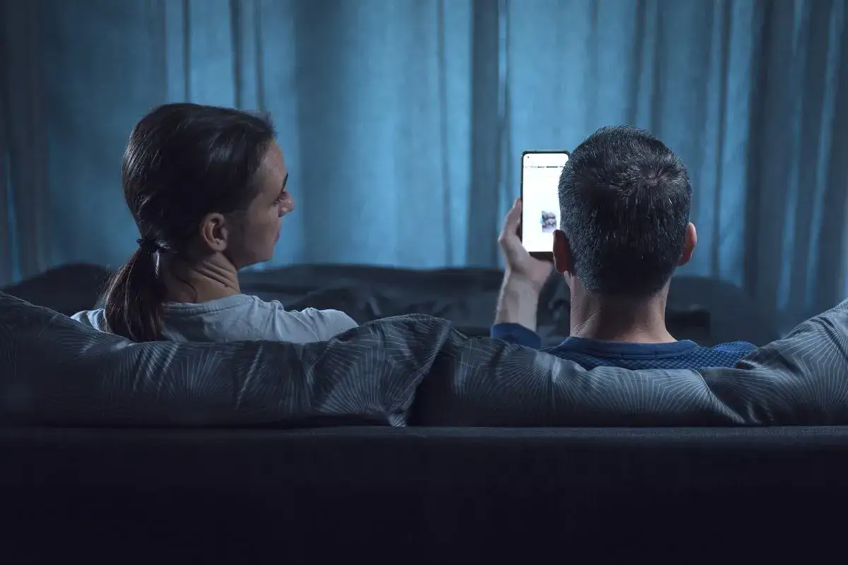 Para siedzi na kanapie, zasłonięta kocem. Kobieta patrzy w bok, mężczyzna trzyma telefon. Nie pisze pierwszy, ale odpisuje.