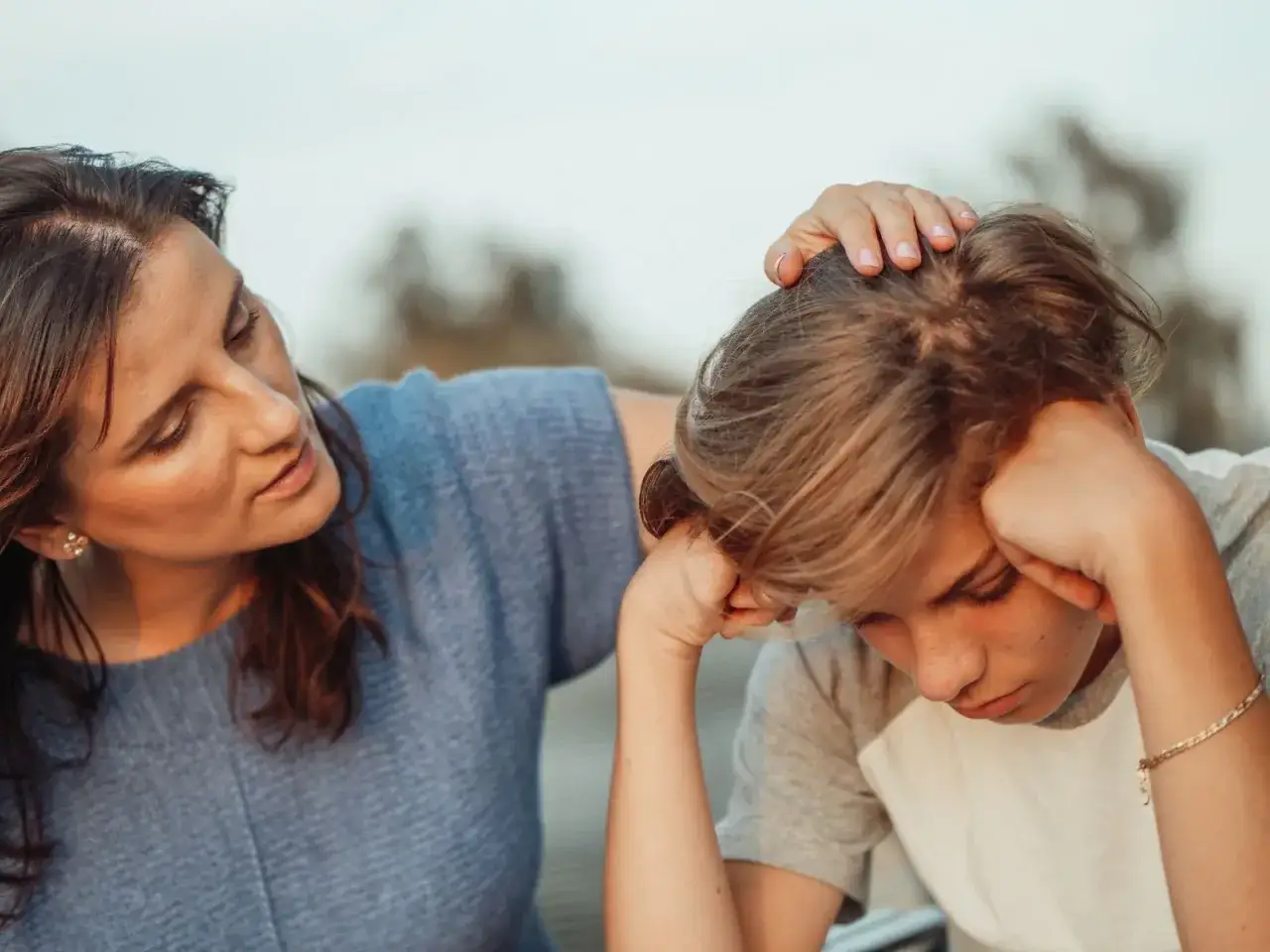 Kobieta pociesza nastolatka, który ma problemy. Jej dłoń spoczywa na jego głowie, okazując wsparcie w trudnym momencie.