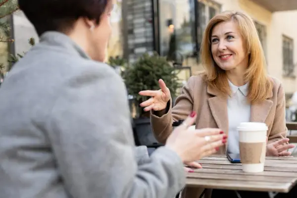 Dwie kobiety rozmawiają przy stoliku, gestykulując. Jedna z nich, z blond włosami, uśmiecha się, opowiadając o swoich przemyśleniach na temat granic w związku.